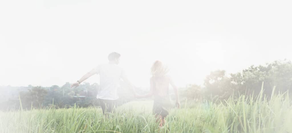 Jeune couple insouciant courant main dans la main dans un champ d'herbe haute et verte sous un ciel lumineux.