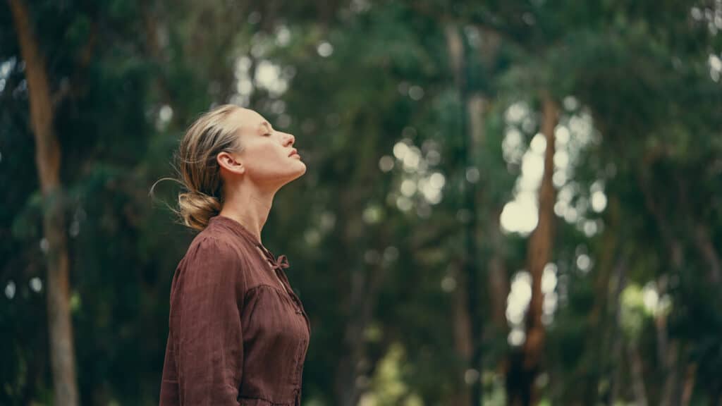 Femme blonde paisible, yeux clos et visage levé, chemise marron, se ressourçant en forêt. Arrière-plan flou de feuillage vert et bokeh.